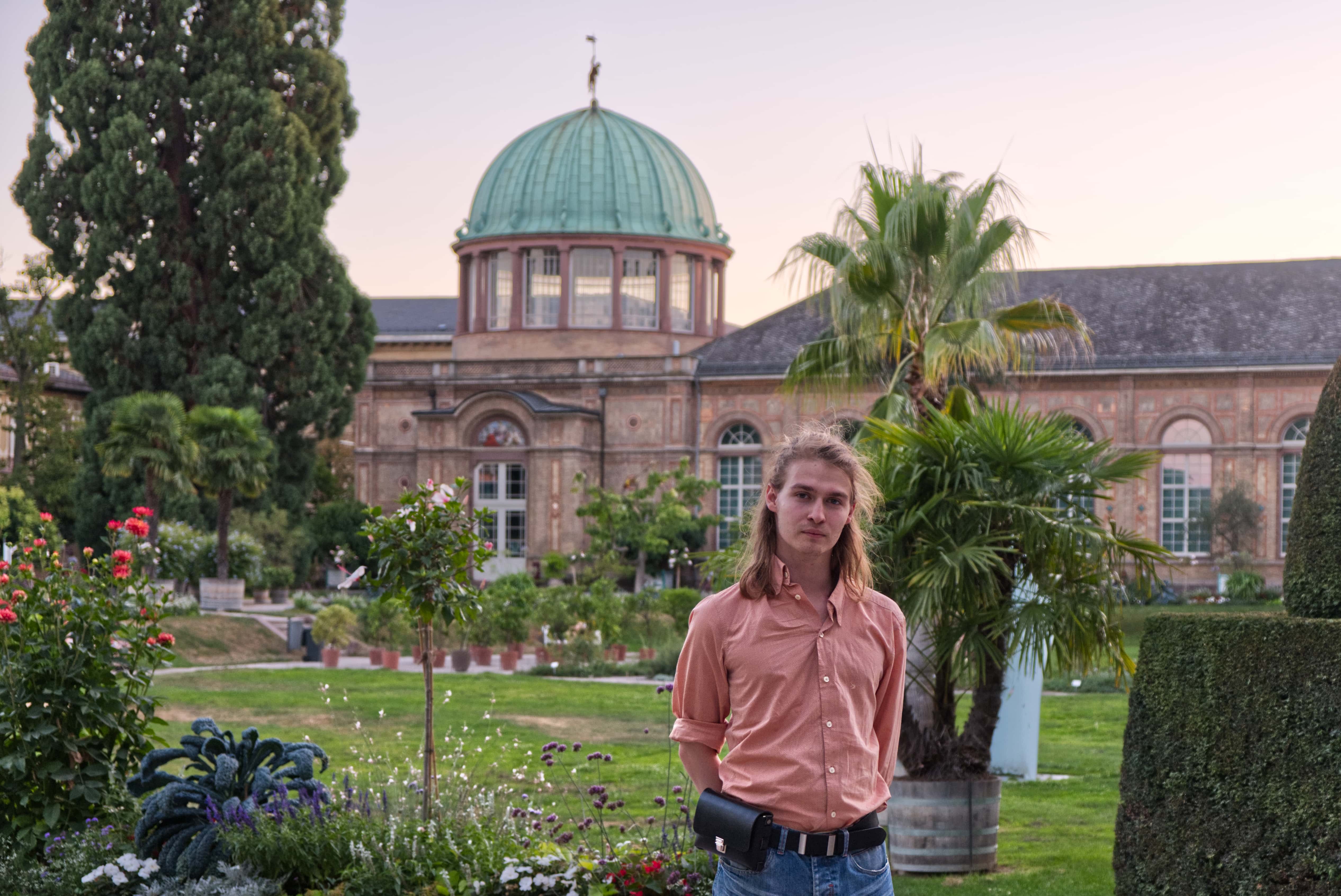 Botanischer Garten und Staatliche Kunsthalle von Karlsruhe – davor steht ein junger mann in rosa Hemd mit schwarzer Gürteltasche und Gürtel
