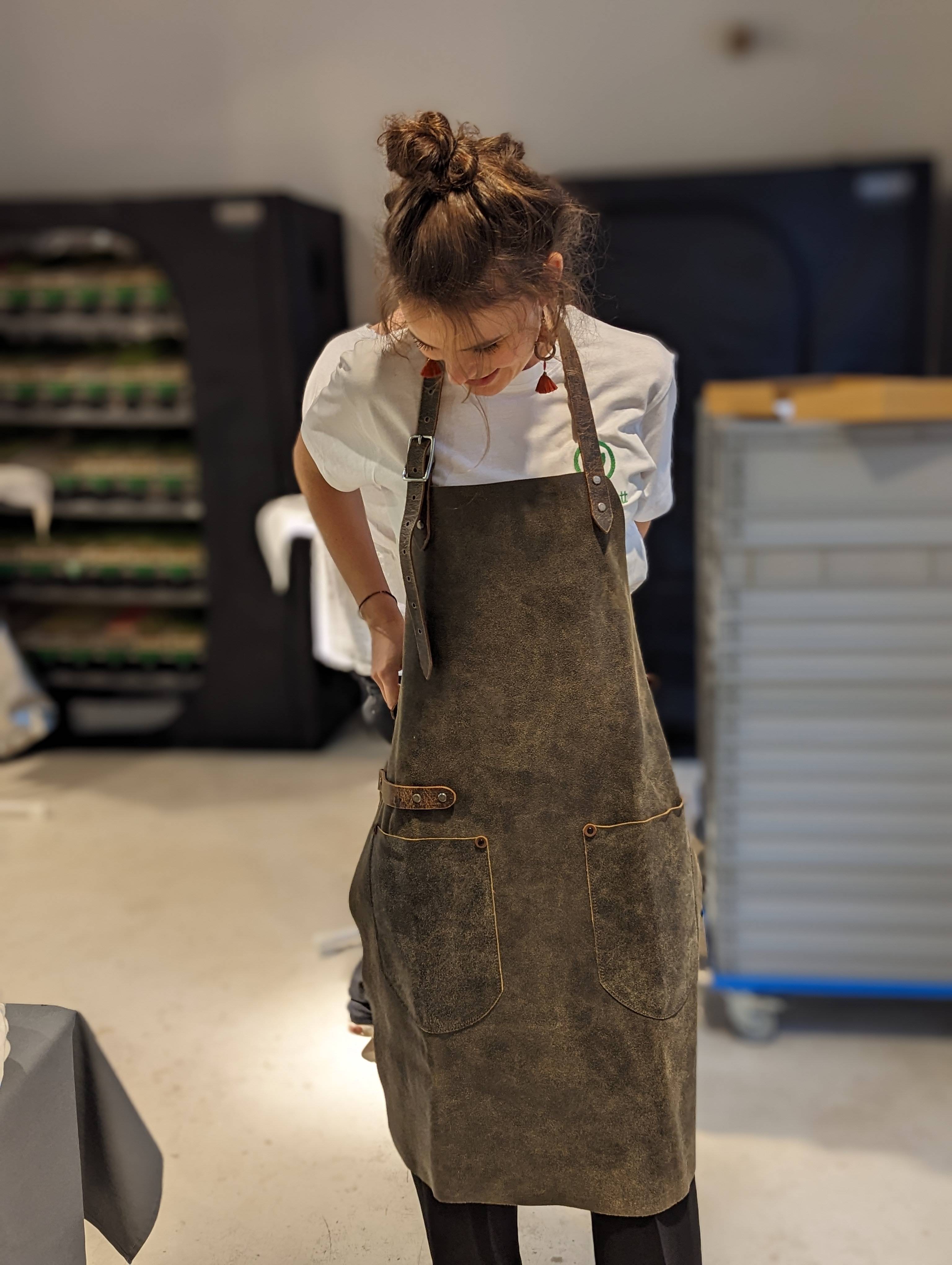 Eine junge Frau trägt eine Lederschürze. Es kann sich um eine Ledergrillschürze oder Lederkochschürze haneln. Sie ist in einem Braunen Wildleder und aus einem Stück Leder. Die Frau steht in einem Innenraum