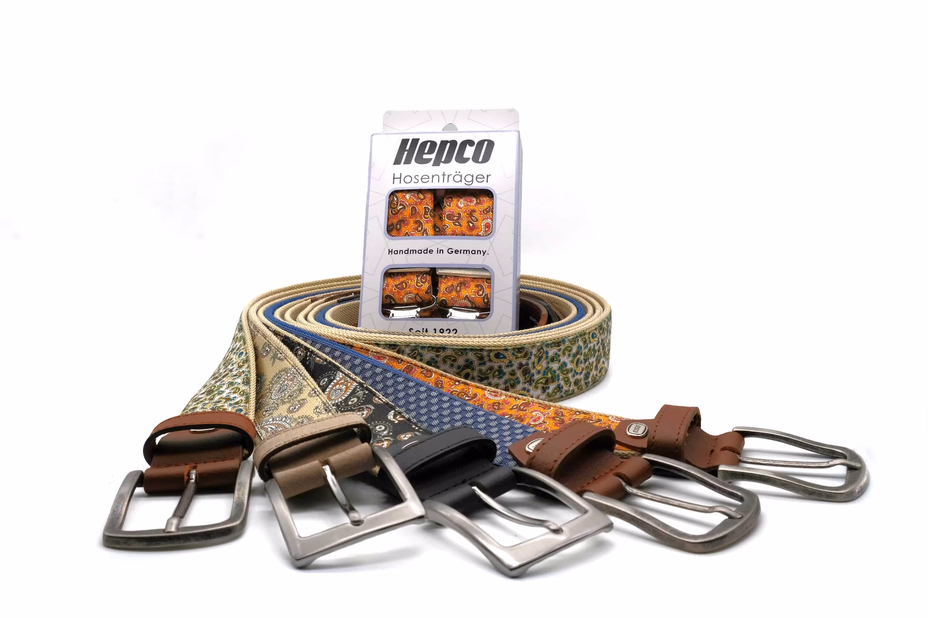 Das Foto zeigt die neuen Hepco-Bandgürtel und Hosenträgerkollektion. Die bunten Gürtel sind gerollt während die schließen vorne präsentiert sind. In der Mitte der Gürtelschnecke befindet sich ein Hepco-Hosenträger mit gleichem Muster wie einer der Bandgürtel.